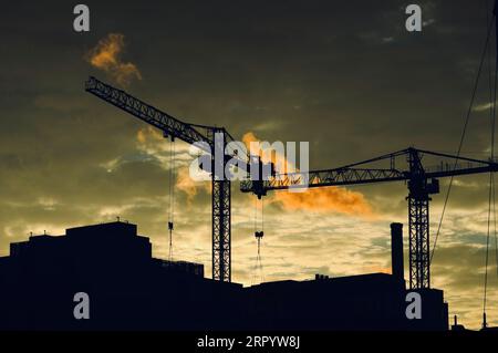 A vibrant sunset illuminates a busy construction site with cranes silhouetted against an orange sky Stock Photo