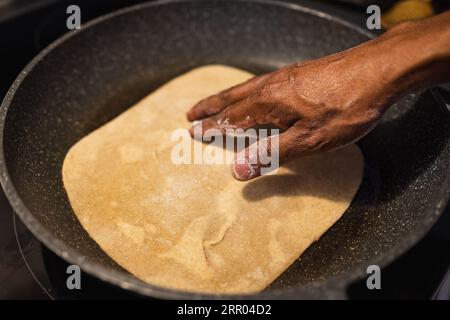 Black man frying flat bread at home. Flat bread with on the frying pan. Fresh multi-grain tortillas. Male hand and homemade naan bread on the pan Stock Photo