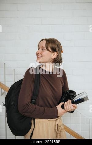 Smiling young woman with backpack and folder looking back while walking in university Stock Photo