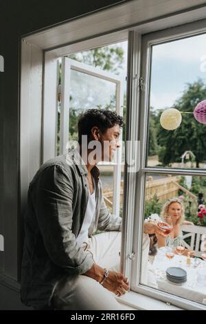 Smiling young man sitting on window sill while looking at friends celebrating party at cafe Stock Photo