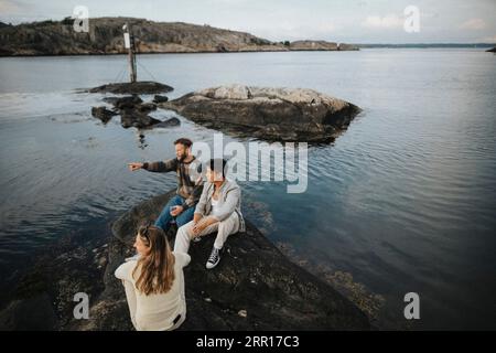 High angle view of man gesturing while sitting with friends on rock near lake Stock Photo