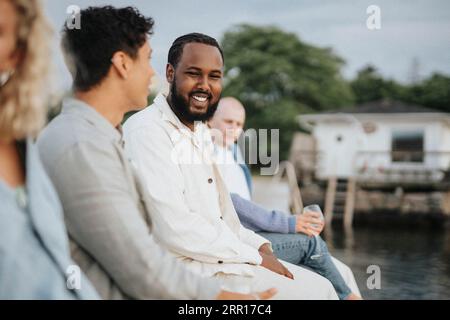 Smiling young man talking with male friend while sitting on pier near lake Stock Photo