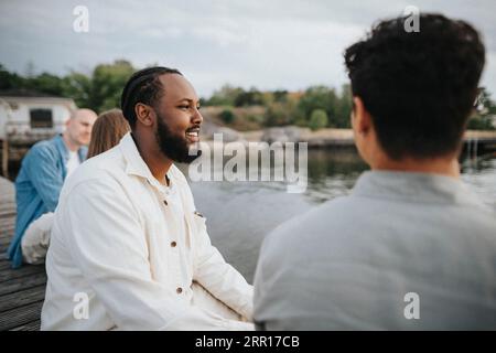 Smiling man looking away while sitting with friend on pier near lake Stock Photo