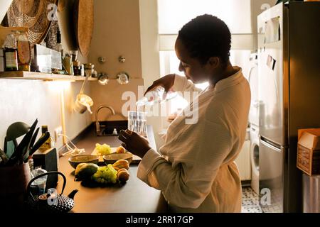 Side view of woman wearing bathrobe pouring water in glass at kitchen counter Stock Photo