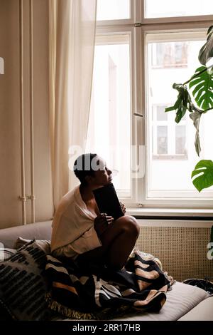 Thoughtful young woman with book sitting on bed at home Stock Photo