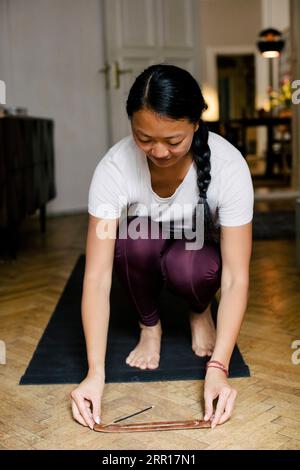 Young woman keeping incense stick on floor while crouching on exercise mat at home Stock Photo