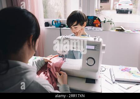 Son looking at mother sewing clothes at home Stock Photo
