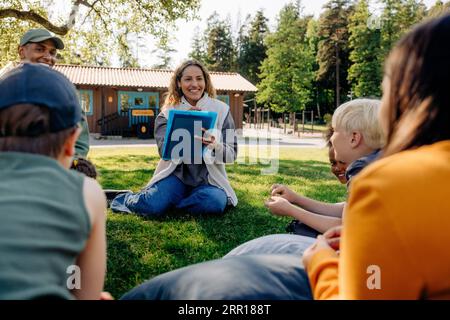 Smiling camp counselors doing activities with children while sitting on grass at summer camp Stock Photo