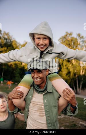 Male camp counselor carrying boy on shoulders while having fun in playground Stock Photo