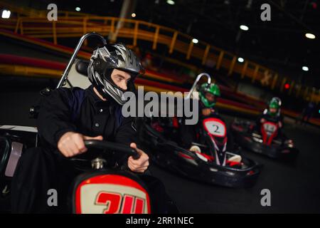 concentrated man driving go kart near diverse drivers in helmets on indoor circuit, adrenaline Stock Photo