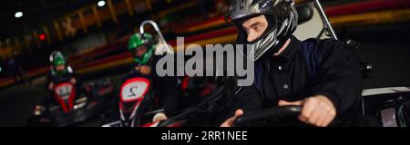 concentrated man driving go kart near diverse drivers in helmets on indoor circuit, banner Stock Photo