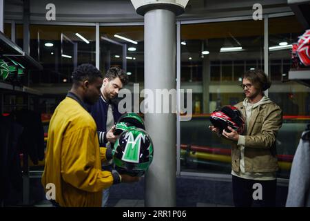 three multicultural friends in casual clothes choosing helmets for karting, go-cart concept Stock Photo
