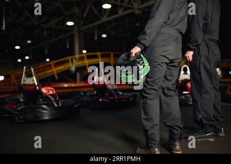 cropped view of two men in sportswear standing on circuit and holding helmets near go kart cars Stock Photo