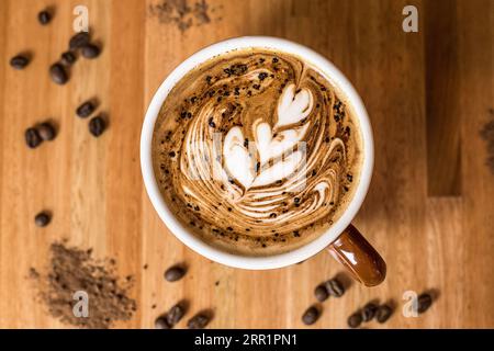 Top view of ceramic mug with fresh aromatic creamy latte coffee placed on wooden dinning table with dry coffee seeds in daylight Stock Photo
