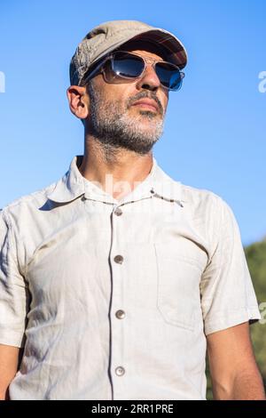 Low angle of confident bearded male wearing white shirt and sunglasses with cap looking away while standing under blue sky Stock Photo