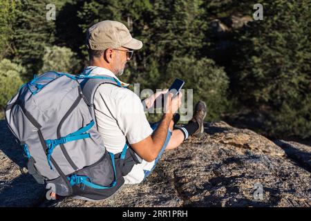 From above bearded male tourist in cap with backpack sitting on edge of rocky cliff above green plants and browsing mobile phone Stock Photo