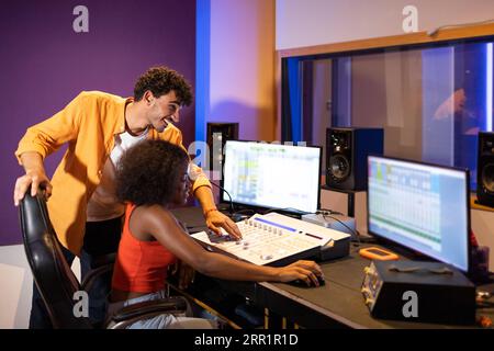Smiling young black female and Hispanic gathered at table with computers and equipment in podcast studio Stock Photo