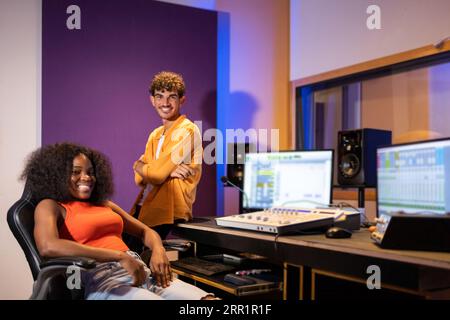 Smiling young black female and crossed hands Hispanic male looking at camera while gathered at table with computers and equipment in podcast studio Stock Photo