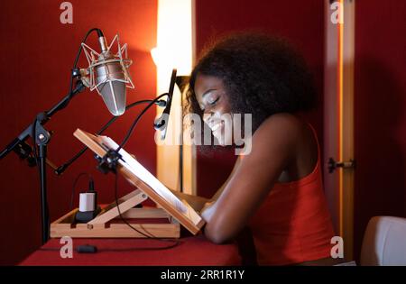 Side view of smiling African American female radio host looking down while sitting at desk with microphone equipment and recording podcast in studio Stock Photo