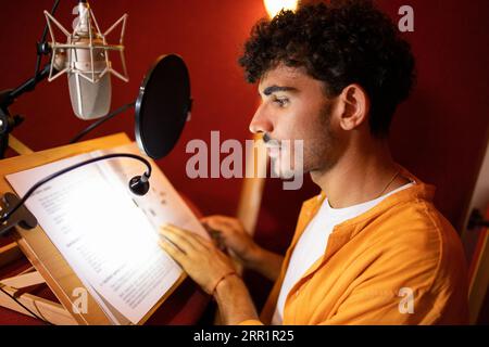 Side view of smiling young Hispanic male radio host looking away while sitting at desk with microphone equipment and recording podcast in studio room Stock Photo