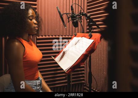 Side view of focused young African American female singer looking down while standing near desk with papers microphone in studio and recording song Stock Photo