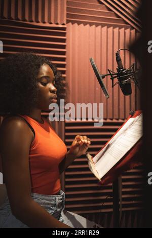 Side view of focused young African American female singer looking down while standing near desk with papers microphone in studio and recording song Stock Photo