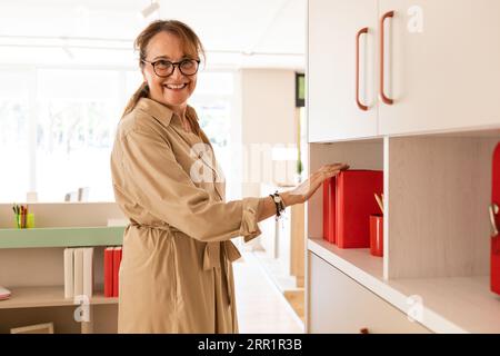 Side view of happy female in casual cloth and accessories standing near cupboard shelf with red ring binder and cups in furniture store looking at cam Stock Photo