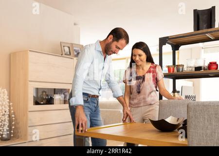 Smiling young couple looking down while standing at sample dining table with chairs and taking measurement with tape in furniture store in daylight Stock Photo