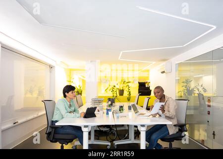 Positive multiracial female colleagues sitting at white desk with laptop tablet and papers and working together in modern office Stock Photo