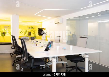 Office meeting room with writing board long table with tablet chairs potted plants on decorated yellow desk glass wall with blurred outside hallway Stock Photo