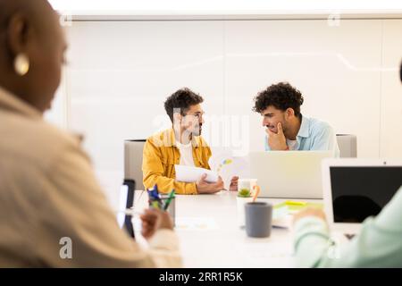 A focused and positive Hispanic man, wearing glasses, sitting at a table with colleagues and working on a laptop with documents Stock Photo