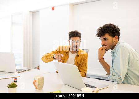 Concentrated positive Hispanic man, wearing glasses, sitting at a table with a colleague and working on a laptop with documents Stock Photo
