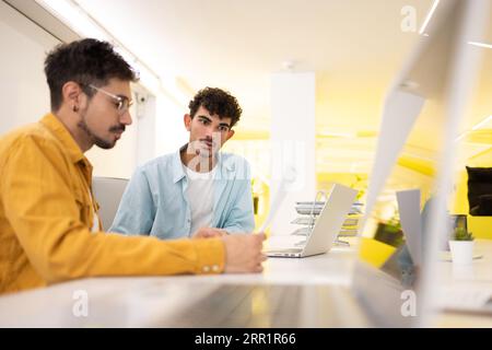 Male colleagues sitting at white desk in modern office and working on project together using laptop and papers Stock Photo