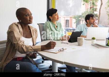 Multiracial coworkers sitting at round desk with laptop tablet and clipboard in modern workplace together and looking away attentively Stock Photo