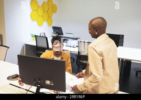 Smiling bearded young male with female while sitting at table with desktop and discussing project details in office lights Stock Photo
