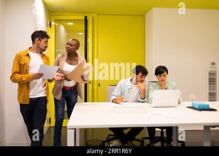 Multiethnic coworkers looking at each other and talking while standing near table with working colleagues looking at screen of laptop in office lights Stock Photo