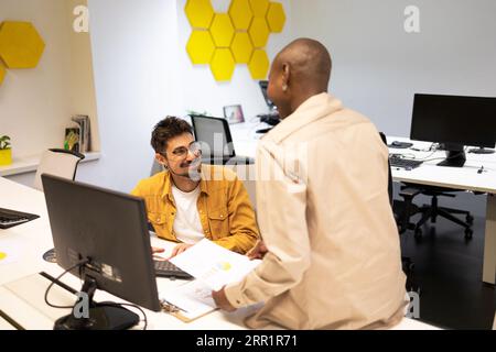 Smiling bearded young male with female looking at each other while sitting at table with desktop and discussing project details in office lights Stock Photo