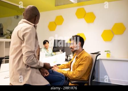 Team of colleagues looking down at documents and working male looking at screen of monitor while sitting at table with ethnic female back view in offi Stock Photo