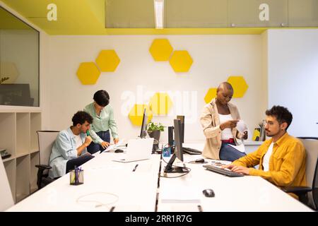 Team of diverse colleagues looking down at documents and working male looking at screen of monitor while sitting at table with ethnic female in office Stock Photo