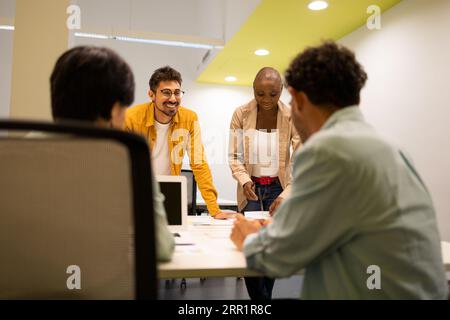 Smiling young man standing with his glasses on with a black woman next to him and two anonymous colleagues sitting at a table talking about a project Stock Photo