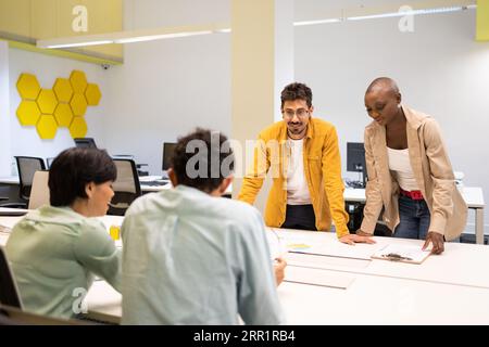 Smiling young man standing with his glasses on with a black woman next to him and two anonymous colleagues sitting at a table talking about a project Stock Photo