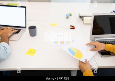 From above of anonymous male looking at screen of laptop while sitting with unrecognizable colleague with printed documents at table in office lights Stock Photo