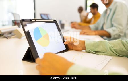 Anonymous male looking at screen of tablet while sitting with unrecognizable colleague with printed documents at table in office lights Stock Photo