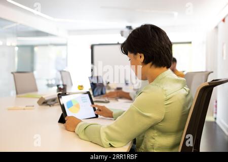 Side view of young female looking at screen of tablet while sitting at table and reading data information of chart picture of project in modern office Stock Photo