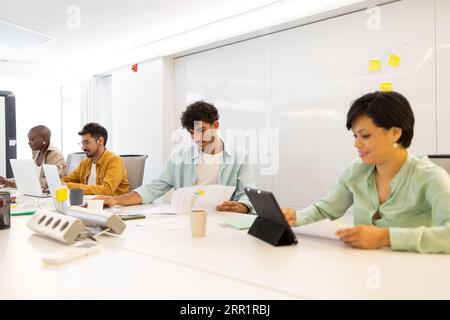 Focused young diverse team members looking at screen of laptop and tablet while sitting working on project with documents gadgets at office table Stock Photo