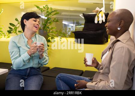 Smiling diverse female colleagues looking at each other while sitting in room with potted plants see through glass and chatting during coffee break Stock Photo