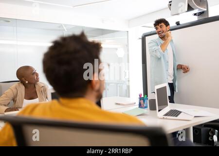 Happy man writing with a marker on a whiteboard while working in a modern office with his colleagues Stock Photo