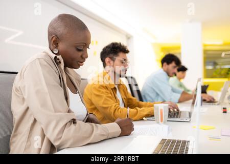 Focused young diverse team members looking at screen of laptop and tablet while sitting working on project with documents gadgets at office table Stock Photo