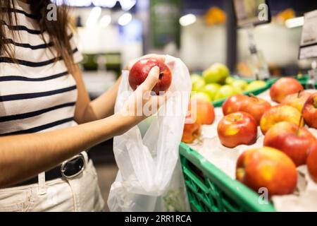 Crop anonymous female customer in casual clothes choosing fresh apples and putting in plastic bag at local market Stock Photo