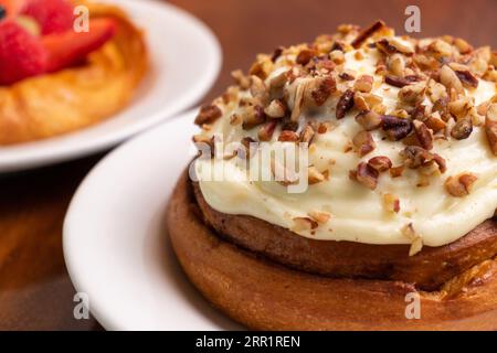 Focus on an appetizing homemade cake with sweet cream and nuts on a white ceramic plate on a wooden dining table with other cakes in a blurred backgro Stock Photo
